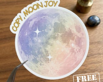 Puede incluir: Una pegatina circular con una luna de estilo acuarela con un degradado pastel de morado, azul, amarillo y rosa. El texto "COPY, MOON JOY" está impreso en dorado en el borde superior y se ven tres estrellas blancas de cuatro puntas.