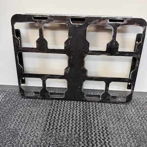 May include: A dark metal rectangular frame with multiple rectangular cutouts. The frame has a slightly weathered appearance, suggesting it may be used for industrial or mechanical purposes. The frame is positioned against a neutral background.
