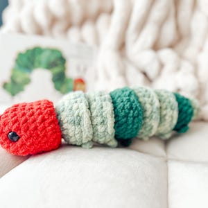 May include: A handmade crochet caterpillar toy in red, green, and light green. The caterpillar has a red head with black eyes and is segmented in shades of green. The toy is placed on a white surface.