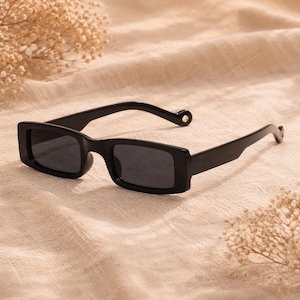 Peut inclure: Lunettes de soleil rectangulaires noires avec des verres foncés. Les lunettes ont une monture noire épaisse et un petit accent doré sur les branches. Les lunettes reposent sur un tissu beige.