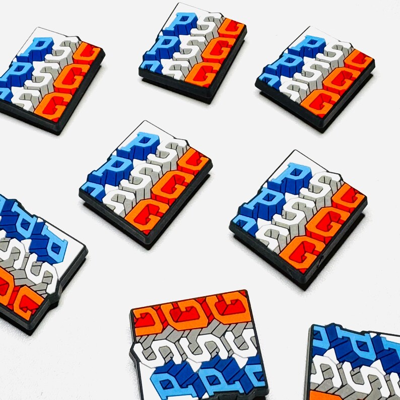 May include: A collection of rectangular charms in various colours. Each charm displays the letters "PSCG" in white, blue, and orange, with a black border. The charms are arranged on a white background.