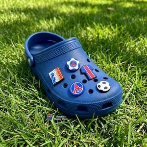 May include: A navy blue clog shoe adorned with football-themed charms, including a football, team emblems, and a jersey design. The shoe rests on green grass. Text on the image reads: Includes a Mystery Charm.