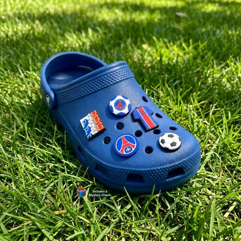 May include: A navy blue clog shoe adorned with football-themed charms, including a football, team emblems, and a jersey design. The shoe rests on green grass. Text on the image reads: Includes a Mystery Charm.
