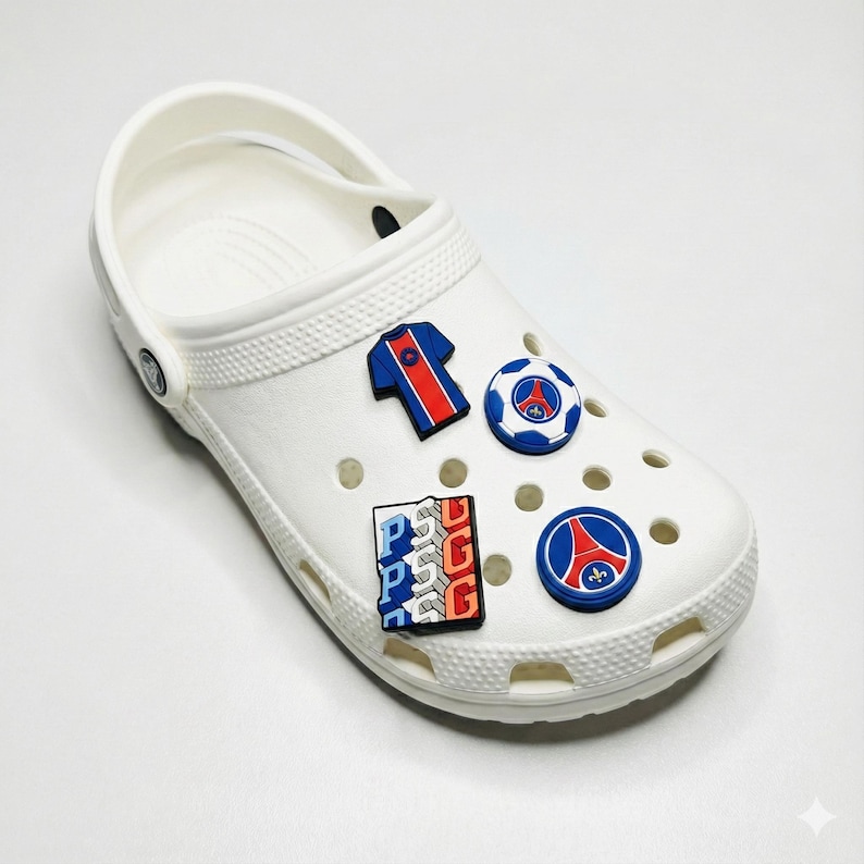 May include: White Crocs shoe decorated with football-themed charms. The charms feature a football shirt, a football, and logos in red, white, and blue. The shoe has multiple holes and a strap.