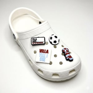 May include: A white clog shoe decorated with four sports-themed charms. The charms include a black and white football, a red and white striped scarf, and two rectangular signs reading Villa Park City of Birmingham and Villa Till I Die.