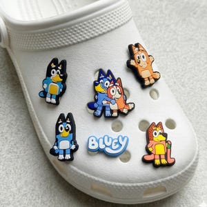 Könnte beinhalten: Weißer Clog-Schuh mit bunten Cartoon-Charms. Die Charms zeigen Charaktere in Blau, Orange und Gelb, mit dem Wort "Bluey" in Blau. Ein lustiges und verspieltes Accessoire für Schuhe.