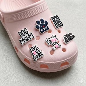 Può includere: Una scarpa zoccolo rosa chiaro adornata con vari ciondoli a tema cani. I ciondoli includono frasi come "Dog Mom", "Dog Dad" e "I Love My Dog", insieme a impronte di zampe e illustrazioni di cani. I ciondoli sono in bianco e nero con accenti rosa.