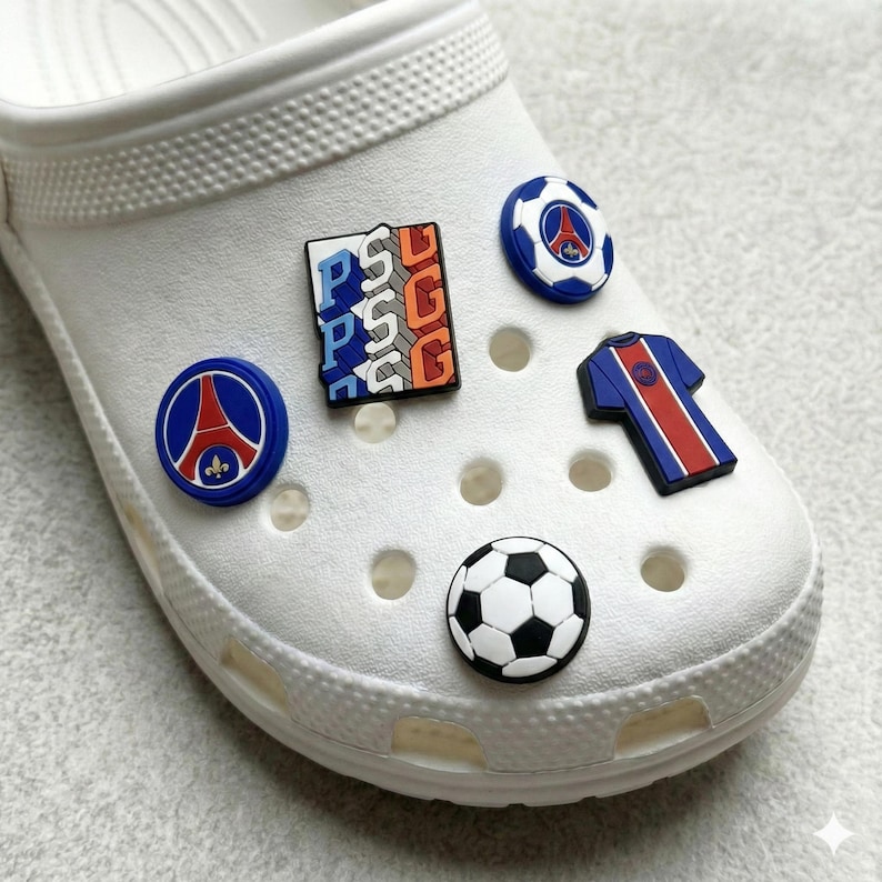May include: White clog shoe adorned with charms. The charms feature a football, a football shirt, and logo designs in blue, red, and white. The charms are attached to the top of the shoe.