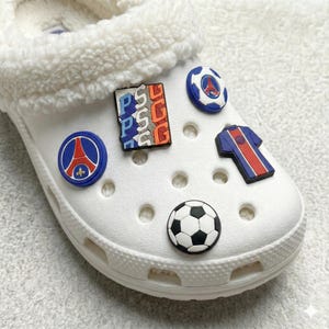 May include: White Crocs shoe with a fluffy white interior, decorated with colourful charms. Charms include a football, a football shirt, and PSG logo designs in blue, red, and white.
