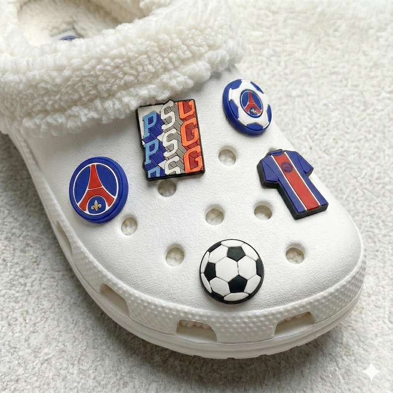 May include: White Crocs shoe with a fluffy white interior, decorated with colourful charms. Charms include a football, a football shirt, and PSG logo designs in blue, red, and white.