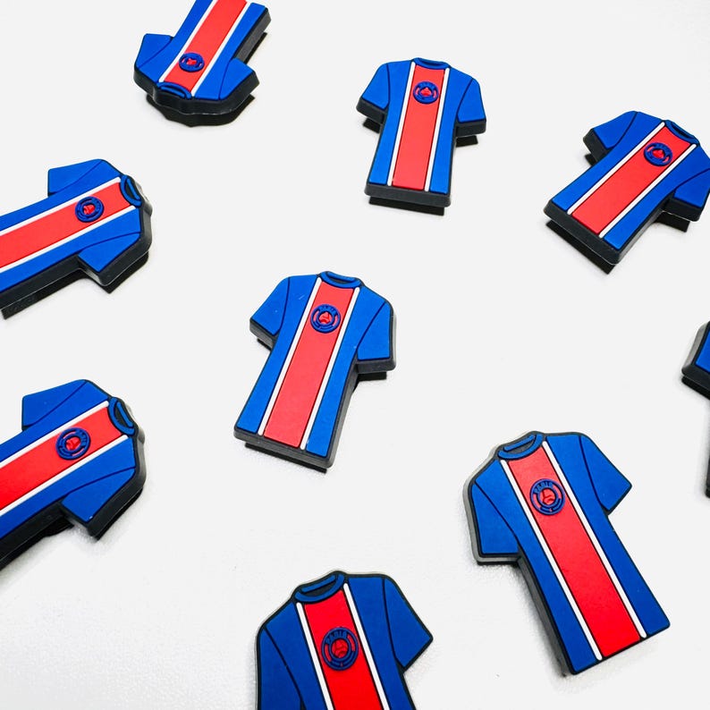 May include: A collection of blue and red football shirt-shaped magnets. Each magnet features a central red stripe and a circular emblem. The magnets are arranged on a white background.