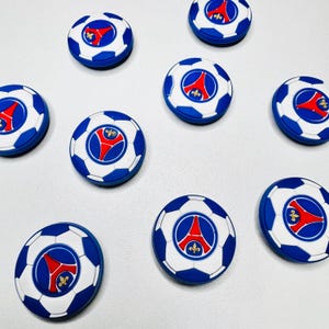 May include: A selection of blue, white, and red soccer ball-shaped magnets. Each magnet features a soccer ball design with a central emblem. The emblem is red with a white outline and a blue border. The magnets are arranged on a white surface.