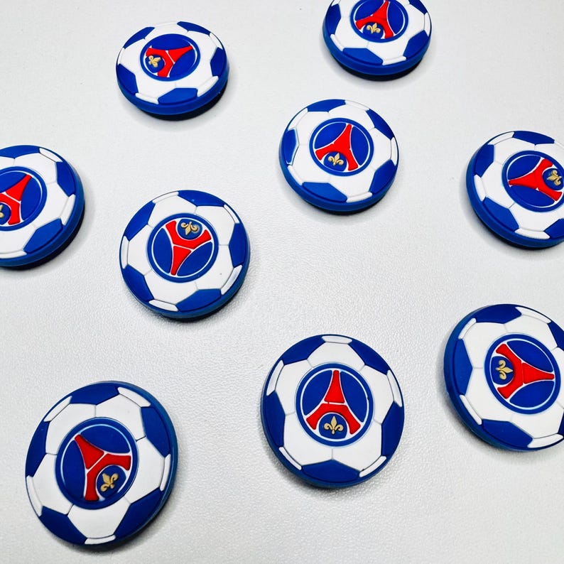 May include: A selection of blue, white, and red soccer ball-shaped magnets. Each magnet features a soccer ball design with a central emblem. The emblem is red with a white outline and a blue border. The magnets are arranged on a white surface.