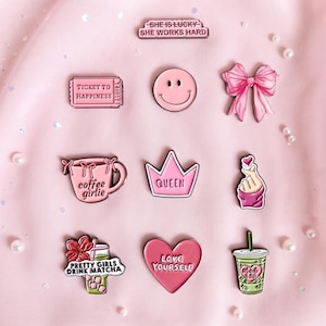 Può includere: Una collezione di nove spille smaltate nei toni del rosa e del verde. I disegni includono un biglietto, una faccina sorridente, un fiocco, una tazza di caffè con testo, una corona, un gesto della mano, una bevanda matcha e un cuore con testo.