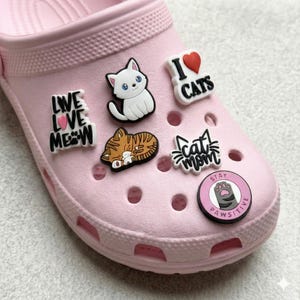 May include: A light pink clog shoe adorned with various cat-themed charms. Charms include a white cat, a sleeping tabby cat, and phrases like "I ❤️ CATS", "Cat Mom", and "Stay Pawsitive".