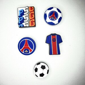 May include: Five colourful charms with a football and Paris Saint-Germain (PSG) theme. Includes a football, a jersey, and various logo designs in blue, red, white, and orange. These charms are likely for shoes or accessories.