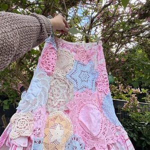 May include: A patchwork skirt with pastel-coloured crochet doilies and fabric patches. The skirt has a pink and white striped fabric base with a ruffled hem. The skirt is held up by a hanger against a backdrop of green foliage.