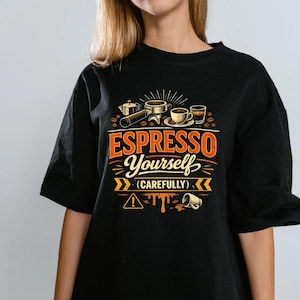 Può includere: T-shirt nero con grafica a tema caffè. Il design include attrezzature per fare il caffè, chicchi di caffè e la scritta "ESPRESSO Yourself (CAREFULLY)" in arancione e bianco. È incluso anche un simbolo di avvertimento.