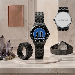 May include: Black wristwatch with a black metal band and a round face featuring the MOPAR logo in blue and white. The watch has a black bezel with white numerals. A matching black ring and two other watches are also visible.