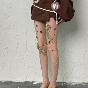 Puede incluir: Medias de rejilla color piel adornadas con adornos florales dorados y rosas y pequeñas estrellas negras. La modelo lleva tacones marrones estampados con un tacón pequeño. El conjunto incluye una minifalda y un top marrones.