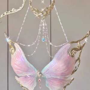 Könnte beinhalten: Ein zartes, flügelförmiges Accessoire in Schmetterlingsform. Die Flügel sind zartrosa mit schillernden Highlights, akzentuiert durch goldfarbene Filigranarbeiten und funkelnde Kristallverzierungen. Ein kleiner, mit Juwelen besetzter Anhänger hängt in der Mitte.