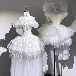 Puede incluir: Dos vestidos blancos sobre maniquíes. El vestido de la izquierda presenta un corpiño con volantes, cintura tipo corsé y una falda en capas con plumas y tul. El vestido de la derecha tiene un tocado de lazo grande, hombros con volantes y una falda voluminosa.