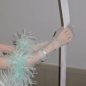 May include: Sheer, light blue gloves with a delicate, feathery trim. The gloves are accented with a silver bracelet. The sleeves are adorned with fluffy, light blue feathers. The gloves are made of a sheer, lightweight material.