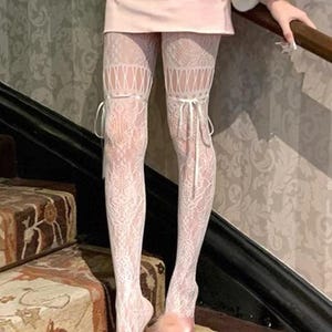 Może przedstawiać: Białe pończochy koronkowe z kwiatowym wzorem i wstążkami. Pończochy są połączone z różowymi satynowymi obcasami z puszystym akcentem. Zdjęcie zrobiono na schodach z wzorzystym dywanem.