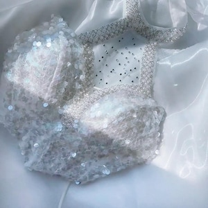 May include: A white, sequined top with a delicate, sheer overlay. The top features a square neckline and is embellished with small, sparkling beads. The fabric appears soft and flowing, creating a glamorous and ethereal look.