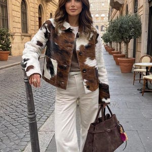 May include: A cowhide jacket with brown, black, and white patches is worn over a neutral-toned outfit. The jacket has a button-up front and a collar. A brown suede handbag is carried, and the person is standing on a cobblestone street.