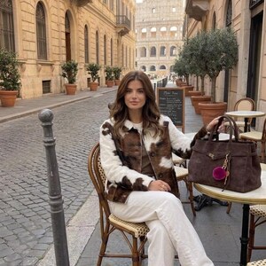 May include: A person seated at a cafe table in Rome, Italy, wearing a brown and white cowhide jacket, white trousers, and tan flats. They hold a brown handbag with a pink pom-pom charm. The Colosseum is visible in the background.