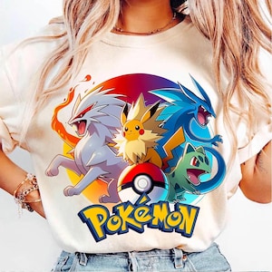 May include: Cream-colored t-shirt featuring a colorful graphic of Pokemon characters, including Pikachu, and a Poke Ball. The word "Pokemon" is written in yellow and blue. The design is set against a circular background with red, orange, and purple hues.