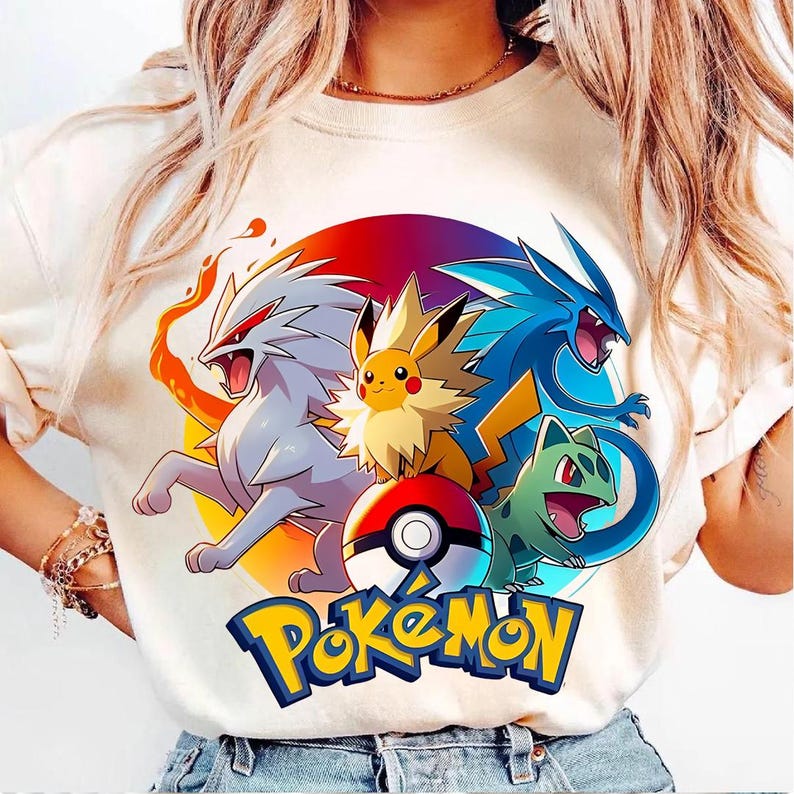 May include: Cream-colored t-shirt featuring a colorful graphic of Pokemon characters, including Pikachu, and a Poke Ball. The word "Pokemon" is written in yellow and blue. The design is set against a circular background with red, orange, and purple hues.
