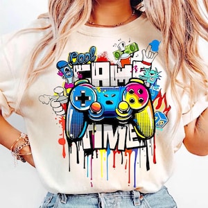May include: Cream-colored t-shirt featuring a colorful graphic of a video game controller with the words "Cool Game Time." The design includes graffiti-style elements, dripping paint, and cartoon characters.