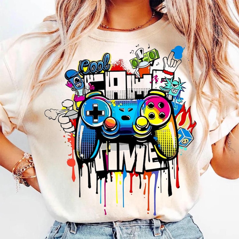 May include: Cream-colored t-shirt featuring a colorful graphic of a video game controller with the words "Cool Game Time." The design includes graffiti-style elements, dripping paint, and cartoon characters.