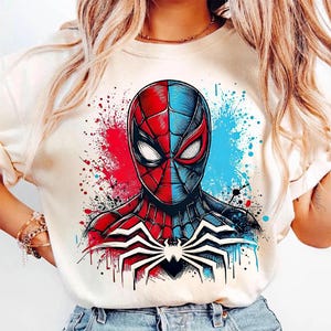 May include: Cream-colored t-shirt featuring a split-face Spider-Man graphic. The design is half red and half blue, with a white spider emblem on the chest. The background has red and blue paint splatters.