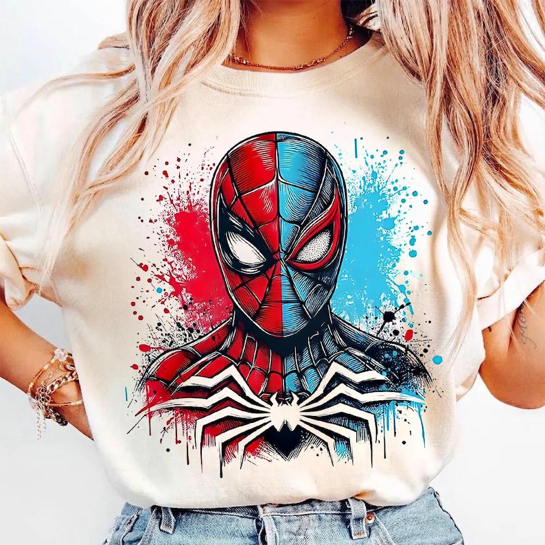 May include: Cream-colored t-shirt featuring a split-face Spider-Man graphic. The design is half red and half blue, with a white spider emblem on the chest. The background has red and blue paint splatters.