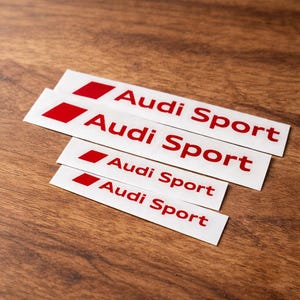Puede incluir: Cuatro pegatinas rectangulares de vinilo blanco de varios tamaños, cada una con un icono de paralelogramo rojo a la izquierda seguido del texto rojo Audi Sport. Las pegatinas están apiladas, mostrando el logotipo de la marca claramente.
