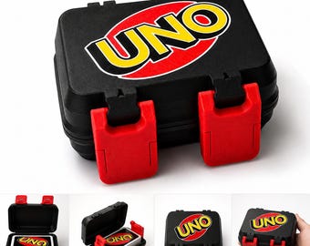 May include: A black rectangular hard-shell carrying case for a card game, featuring the yellow and red UNO logo on the lid. The case has two bright red plastic latches on the front. The interior holds a deck of playing cards with matching branding.