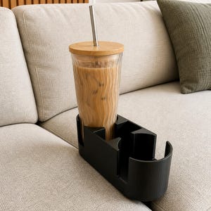 Op de afbeelding: Een glas ijscoffie met een bamboe deksel en metalen rietje staat in een zwarte bekerhouder. Het glas heeft een textuurontwerp. De bekerhouder staat op een lichtgekleurde bank, met een groen kussen op de achtergrond.