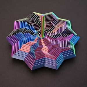 Op de afbeelding: Een meerlagige, stervormige sculptuur met een levendige iriserende afwerking. Het kunstwerk toont een spectrum aan kleuren, waaronder roze, blauw en groen, tegen een donkere achtergrond. Een verticale, metalen staaf vormt het centrale element.