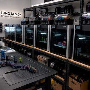 May include: A 3D printing workshop featuring a row of Bambu Lab printers on metal shelving, with filament spools above. A black table in the foreground displays various small 3D printed figures and a long articulated dragon. A sign reads LUNQ DESIGN.