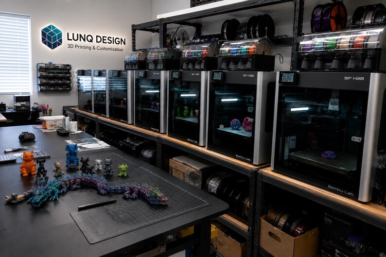 May include: A 3D printing workshop featuring a row of Bambu Lab printers on metal shelving, with filament spools above. A black table in the foreground displays various small 3D printed figures and a long articulated dragon. A sign reads LUNQ DESIGN.