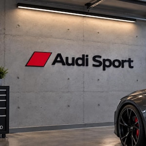 Può includere: Un'insegna da parete Audi Sport con un logo a parallelogramma rosso accanto al testo nero Audi Sport. L'insegna è esposta su una parete in cemento grigio all'interno di un garage con una vista parziale di un'auto grigio scuro e un mobiletto.