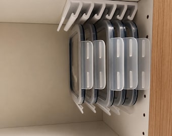 May include: A white plastic under-shelf storage rack mounted to the top of a cabinet, holding four rectangular food storage lids in a vertical, hanging position. The lids are clear with translucent white locking tabs on the side edges.
