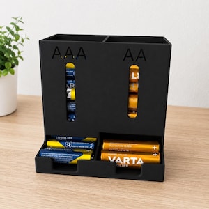 Op de afbeelding: Een matzwarte kunststof batterijhouder met twee verticale opbergvakken gelabeld AAA en AA. De unit is voorzien van verticale kijkvensters voor het batterijniveau en twee horizontale uitgiftebakjes aan de onderkant met blauwe en gele batterijen.