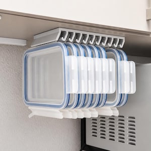 Op de afbeelding: Een lichtgrijs opbergrek voor onder een kastje met zes rechthoekige, doorzichtige plastic bewaarbakjes met blauwe siliconen afdichtingen en witte sluitclips, opgehangen aan een rail onder een keukenkastplank.