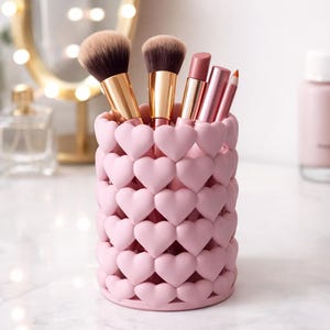 Op de afbeelding: Een cilindrische make-up kwastenhouder met een herhalend patroon van verhoogde, lichtroze hartjes. De houder bevat twee make-up kwasten met gouden hulzen en bruine haren, samen met drie lipproducten in metallic roze hulzen.
