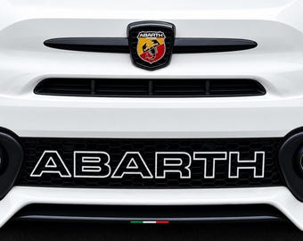 May include: The front bumper of a white Abarth vehicle featuring a black grille with the word ABARTH in white lettering. A black shield emblem with a red, yellow, and black scorpion logo is centered above the grille, flanked by two circular fog lights.