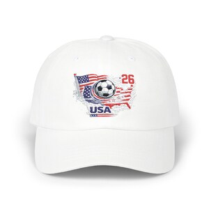May include: White baseball cap featuring a soccer ball graphic inside a USA map design. The design incorporates the American flag colors of red, white, and blue, with the number 26 in red. The word "USA" is also included.